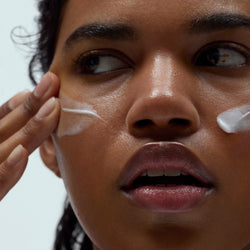 Close-up of a woman applying Dieux's Skin Mercy Recovery Cream to her face with a neutral background