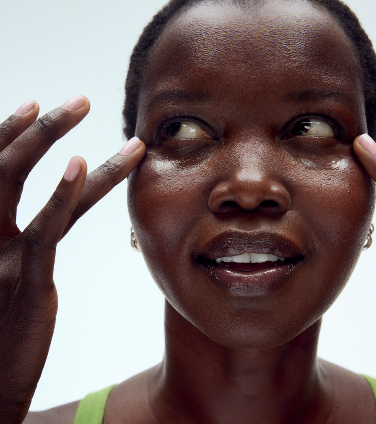 Close-up of a person with deep skin tone gently tapping TEST - Winged Forever Eye Mask under their eyes, smiling softly, in a bright green top against a white background.