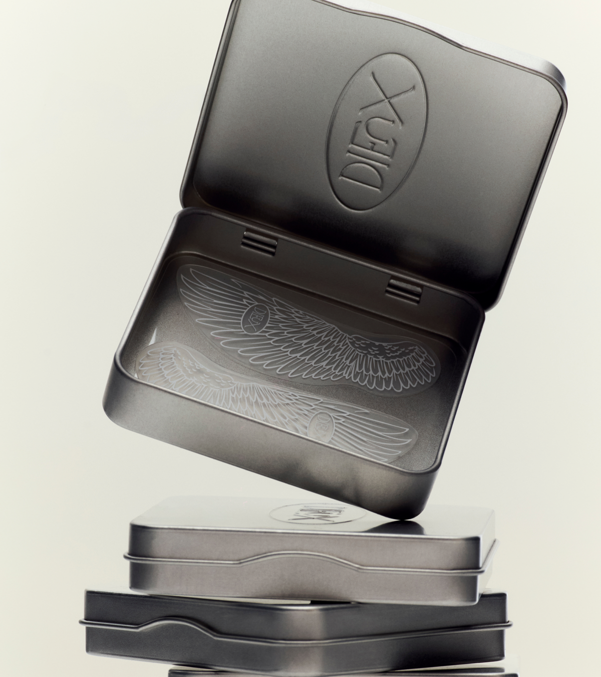 A stack of metallic tins, with the top tin open to reveal an engraved wing design and TEST - Winged Forever Eye Mask embossed on the lid.