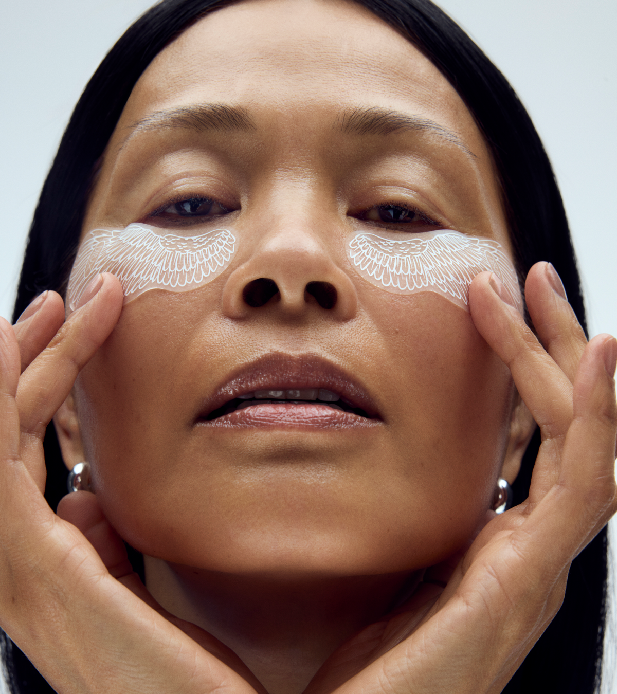 A person with smooth skin applies TEST - Winged Forever Eye Mask under their eyes, gently touching their face. The plain, light background highlights this skincare routine.