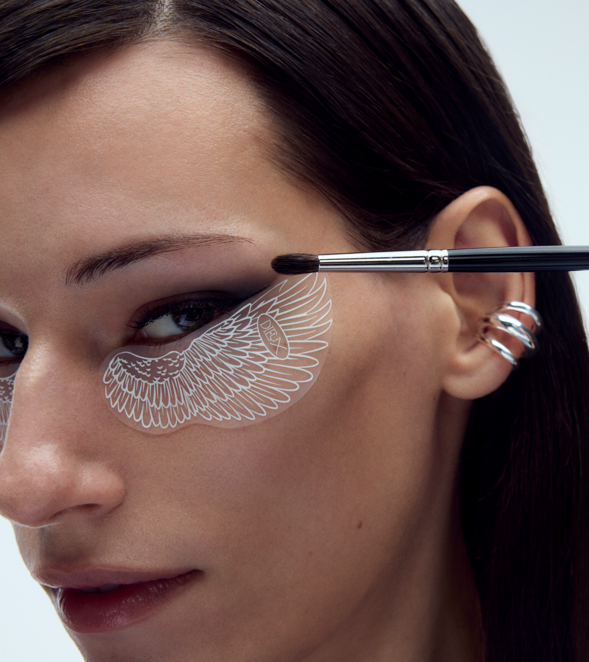 A woman with straight brown hair wears the TEST - Winged Forever Eye Mask under her eyes and applies dark eyeshadow with a brush. She accessorizes with silver ear cuffs.