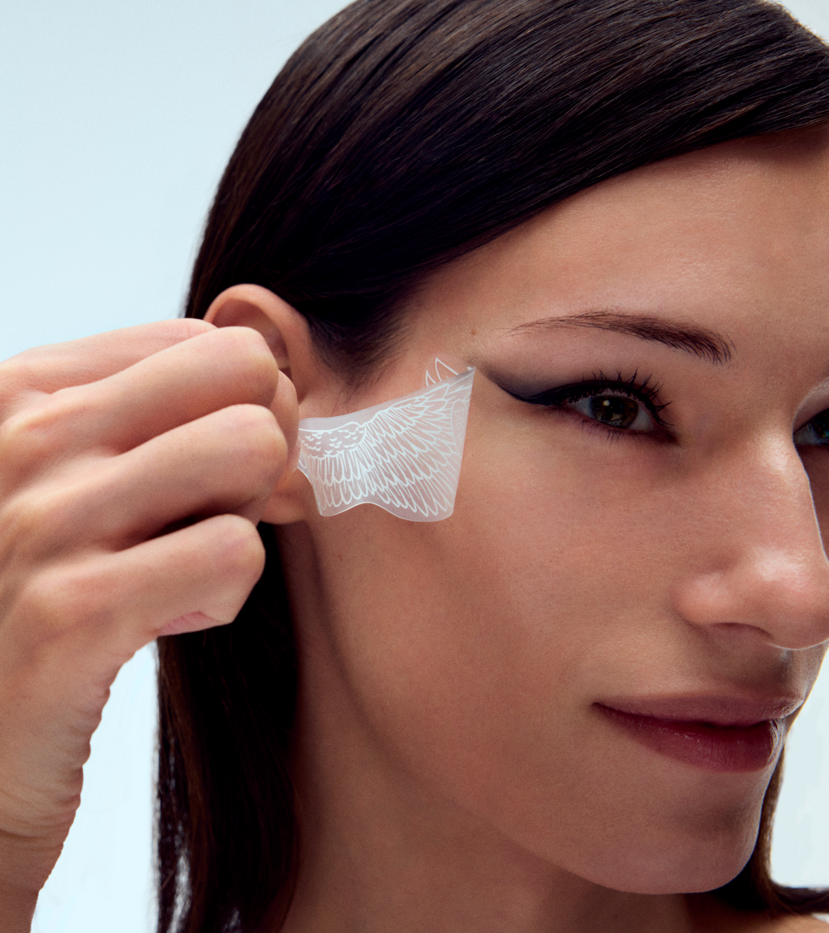 A woman peels off a transparent stencil from beside her eye, revealing sharp black winged eyeliner created with the TEST - Winged Forever Eye Mask.