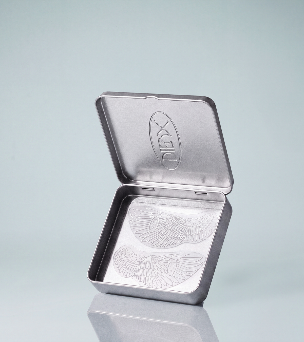 The TEST - Winged Forever Eye Mask comes in a silver metal tin with an embossed logo inside the lid and two wing illustrations at the base, all set on a light blue background.